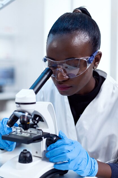 Perto de químico de etnia africana, fazendo análise de amostra, olhando através do microscópio. Cientista negro da saúde no laboratório de bioquímica, usando equipamento esterilizado.