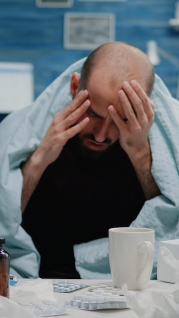 Perto de um adulto doente com dor de cabeça esfregando as têmporas no cobertor. Homem com enxaqueca e sintomas de gripe tentando curar a dor enquanto toma medicamentos, comprimidos, chá e termômetro na mesa