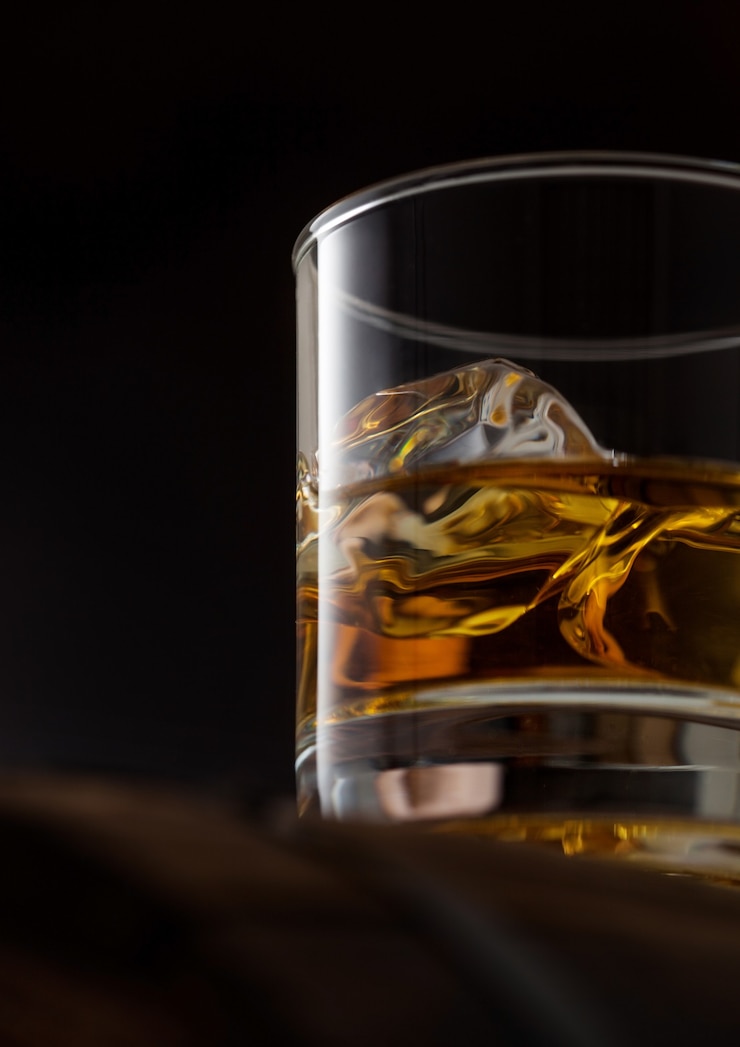 Foto perto de um copo de whisky de single malte com cubas de gelo em cima de um barril de madeira em fundo preto