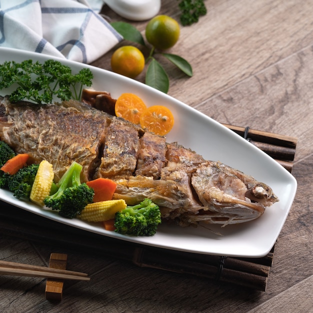 Perto de um delicioso prato de peixe frito em um prato branco com vegetais em uma mesa de madeira