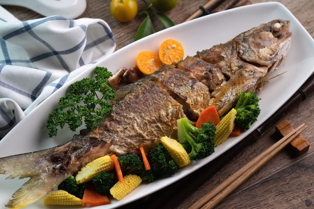 Perto de um delicioso prato de peixe frito em um prato branco com vegetais em uma mesa de madeira