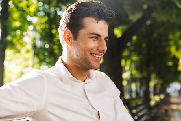 Perto de um homem bonito e sorridente vestido com uma camisa, sentado em um banco no parque