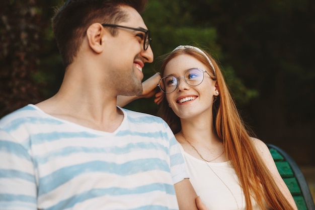 Perto de um jovem casal encantador se divertindo rindo enquanto está sentado em um banco e olhando um para o outro.