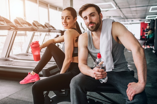 Perto de um lindo casal sentado no banco de esporte e posando. Eles também estão bebendo água de suas garrafas e descansando após um treino duro.