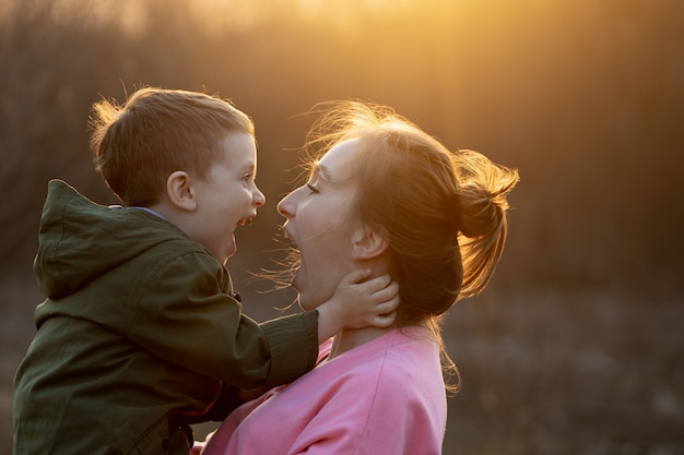 Perto de uma adorável mãe e filho se divertindo ao ar livre. garotinho bonito holded por sua mãe nos braços, que está rindo contra o pôr do sol. conceito de dia das mães