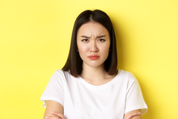 Perto de uma jovem mulher asiática olhando com raiva ou decepcionada, carrancudo descontente para a câmera, em pé na camiseta branca, amarela.