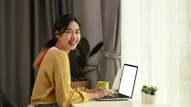 Perto de uma jovem mulher digitando no laptop enquanto está sentada em frente a uma tela em branco do laptop