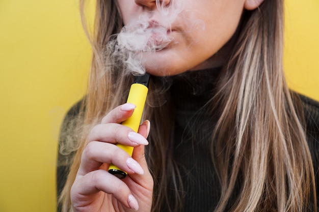 Foto perto de uma jovem que fuma um cigarro eletrônico descartável e exala fumaça. fundo amarelo brilhante. alternativa para cigarros regulares e vapor