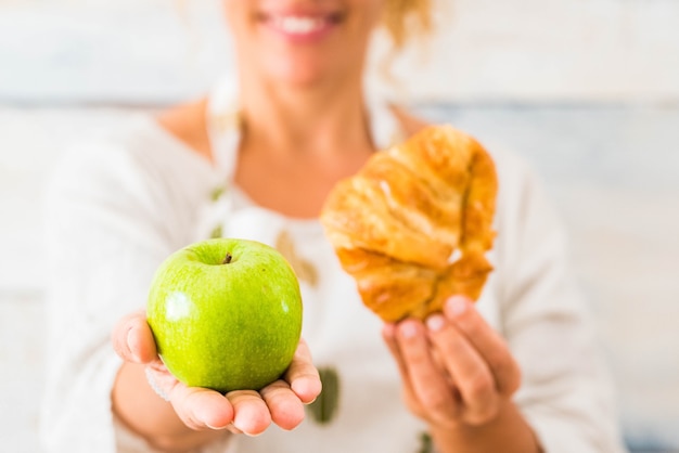 Perto de uma linda mulher segurando uma maçã e um croissant na frente da câmera, escolhendo entre uma vida saudável ou um estilo de vida pouco saudável - fazer dieta e cuidar da comida