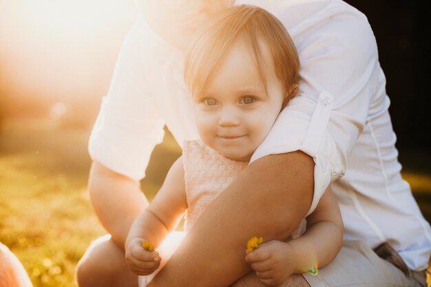 Perto de uma menina olhando para a câmera, sorrindo e segurando uma flor enquanto é abraçada por seu pai contra o pôr do sol.