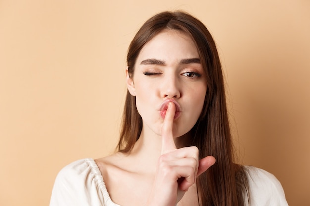 Foto perto de uma mulher atraente silenciando para a câmera, escondendo o segredo, fazendo sinal de silêncio e piscando, mostrar sinal de tabu, de pé sobre fundo bege.