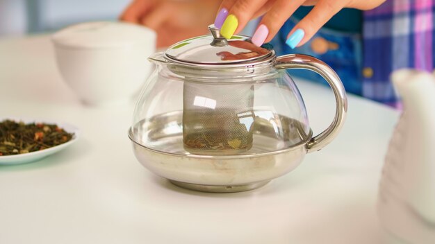 Perto de uma mulher fazendo chá verde pela manhã, no café da manhã, em uma cozinha moderna usando um bule sentado perto da mesa. Ela está colocando com as mãos, pressionando ervas, saudável, folhas de chá, na panela