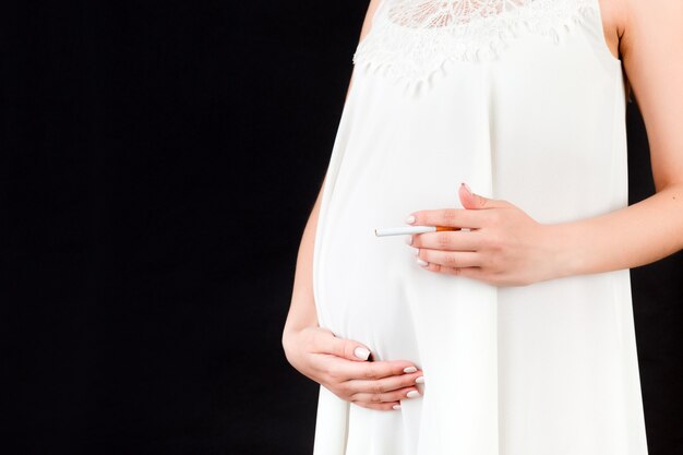 Perto de uma mulher grávida em um vestido branco com um cigarro contra sua barriga em fundo preto. Risco perigoso para o feto.