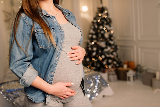 Perto de uma mulher grávida sorridente e feliz tocando sua barriga sobre o fundo da árvore de natal