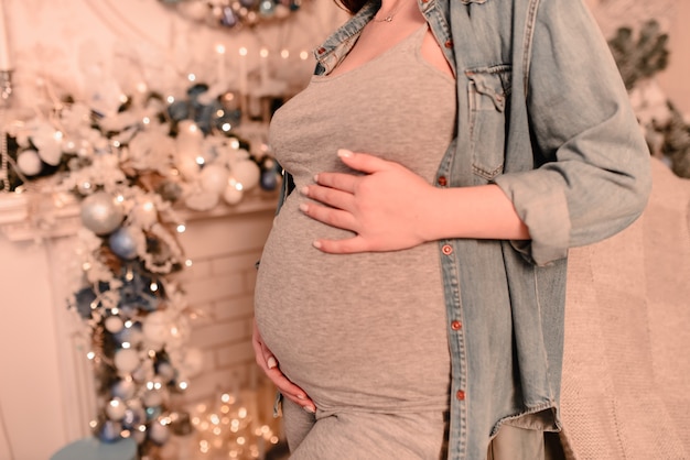 Perto de uma mulher grávida sorridente e feliz tocando sua barriga sobre um fundo de árvore de Natal