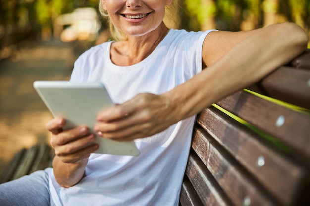 Foto perto de uma senhora alegre segurando um pc eletrônico e sorrindo enquanto está sentada no banco ao ar livre