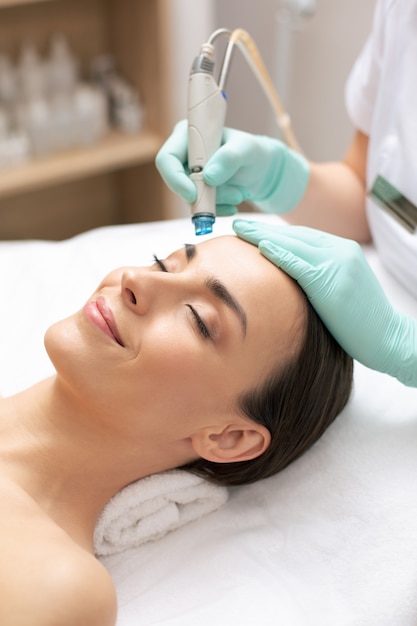 Perto de uma senhora calma e relaxada deitada com os olhos fechados e um cosmetologista profissional limpando sua pele com ferramenta de dermoabrasão