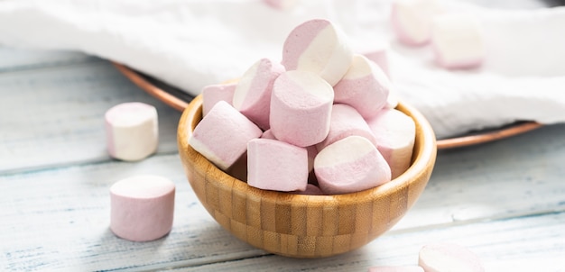 Perto de uma tigela de madeira cheia de marshmallows rosa e brancos com alguns espalhados em uma toalha de mesa branca, bandeja escura e mesa de madeira branca.