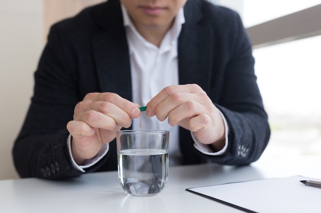 Foto perto do empresário no escritório segurando comprimidos e um copo de água doce, tomando remédio para dor de cabeça, dor abdominal ou tomando vitaminas, conceito de saúde