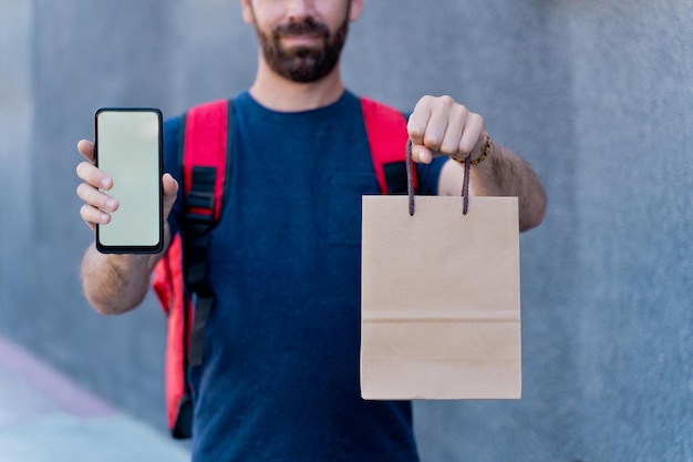 Perto do entregador com maquete de tela do smartphone, entregando a caixa do pacote. Conceito de tecnologia e serviço de correio. Visão horizontal de um piloto irreconhecível entregando um pacote para casa.