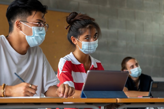 Perto do estudante adolescente menina asiática em sala de aula usando uma máscara facial usando o tablet. Colegas de classe multirraciais. Conceito de educação. Conceito de saúde.