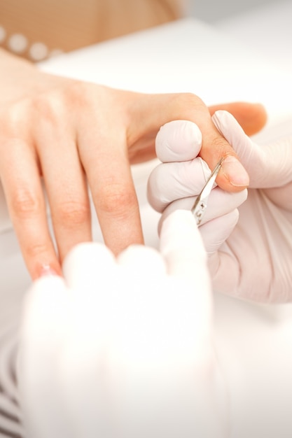 Perto do mestre de manicure com uma tesoura de manicure remove cutículas de unhas femininas no salão de beleza.