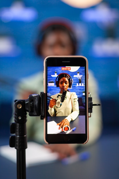 Foto perto do vlog de gravação de smartphone do influenciador africano em estúdio caseiro. falando durante a transmissão ao vivo, blogueiro discutindo no podcast usando fones de ouvido.