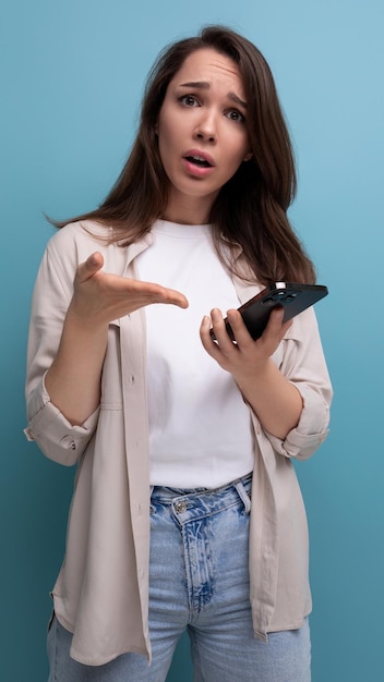 Perturbada linda morena jovem morena vestida com uma camisa e jeans segurando um telefone nas mãos