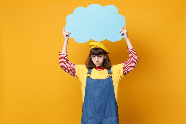 Perturbado adolescente menina perplexa na boina francesa, vestido jeans segurar azul vazio em branco Diga nuvem, balão isolado no fundo amarelo. Conceito de estilo de vida de emoções sinceras de pessoas. Mock up espaço de cópia.