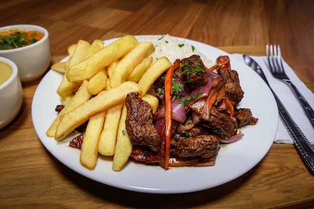 Peruanisches Essen Lomo Saltado, Pommes Frites mit Fleisch, Zwiebeln, gelbem Pfeffer, Petersilie, weißem Reis und Sojasauce, auf einem weißen Teller mit Besteck und Holzhintergrund.