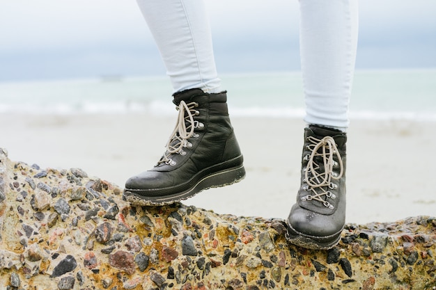 Pés femininos em jeans e botas de inverno em pé sobre uma pedra na costa close-up