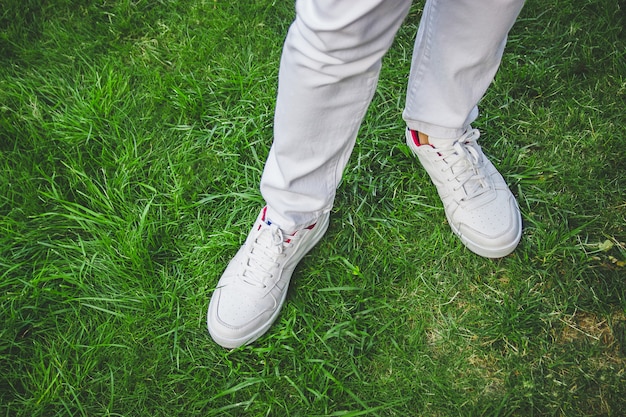 Pés masculinos em sapatos brancos na grama verde. Jeans branco.