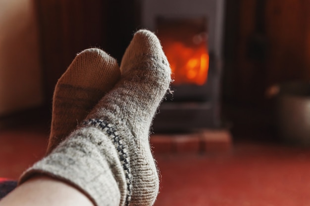 Pés pernas em roupas de inverno meias de lã no fundo da lareira. Mulher sentada em casa na noite de inverno ou outono, relaxante e aquecimento. Conceito de inverno e frio. Véspera de Natal de Hygge.