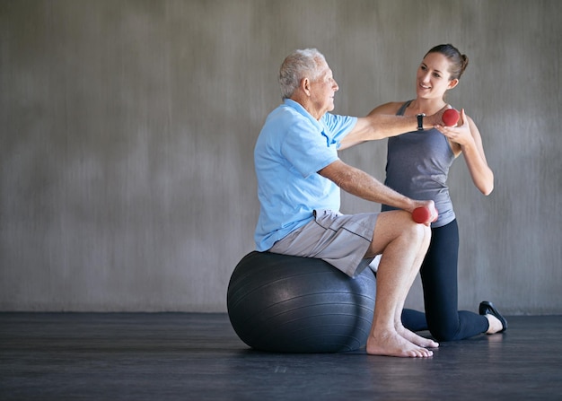 Las pesas del fisioterapeuta y la sonrisa del anciano en la pelota para la aptitud o la rehabilitación en el gimnasio en la maqueta de la persona mayor levantamiento de pesas y entrenador personal ayuda para la salud del cuerpo o la fisioterapia