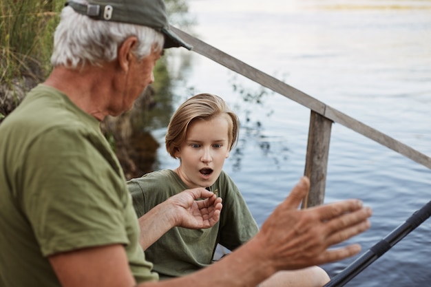 Pesca exitosa, papá habla sobre peces grandes para atrapar durante el fin de semana de verano, pesca con mosca hombre maduro, pescador con nieto, hijo mirando al padre con la boca abierta y expresión facial asombrada