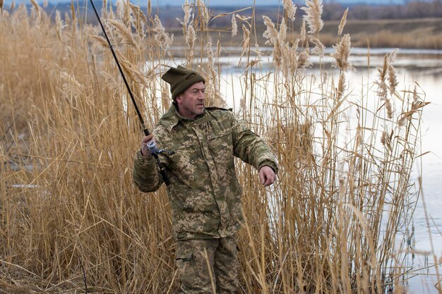 Pesca nos juncos para girar em águas tranquilas Um homem com roupas de camuflagem em clima frio está pescando na margem do rio