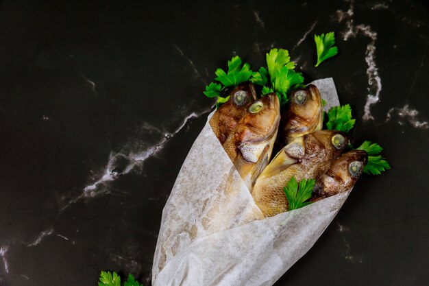 Pescado entero crudo con perejil envuelto en papel pergamino sobre mesa negra.