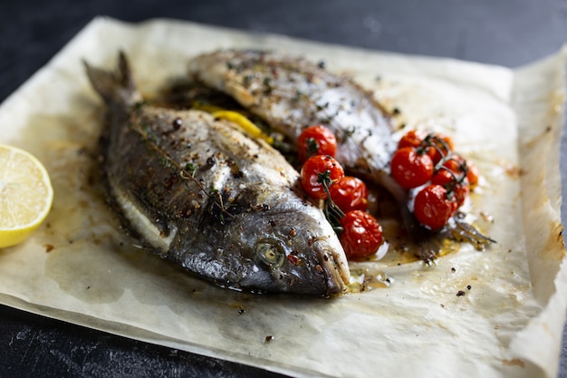 Pescado Fried Dorado en papel con limón, romero y tomates sobre un fondo gris, vista superior. Pescado dorado a la parrilla sobre una placa de piedra.