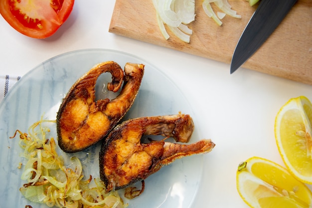 El pescado frito con cebollas está en un plato sobre una toalla de gofres al lado hay una tabla de cortar con picada en ...