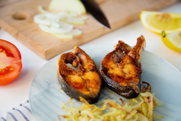 El pescado frito con cebollas está en un plato sobre una toalla de gofres al lado hay una tabla de cortar con picada en ...