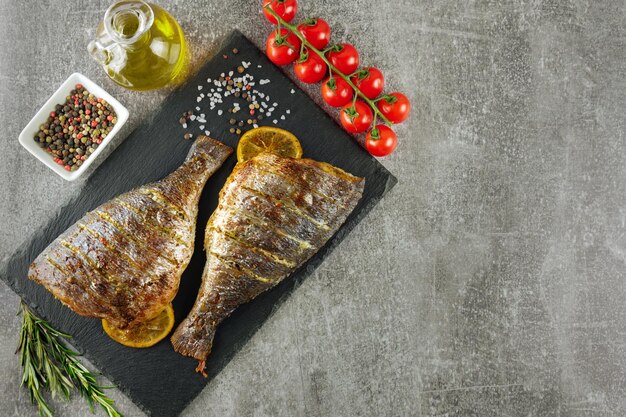 Pescado a la parrilla asado con limón, romero, tomates, aceite de oliva y especias en un plato de pizarra negra