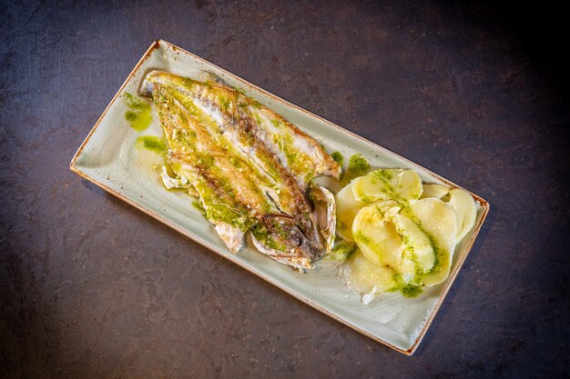 Pescado en salsa verde con patatas sobre un fondo negro, sobre una placa blanca.