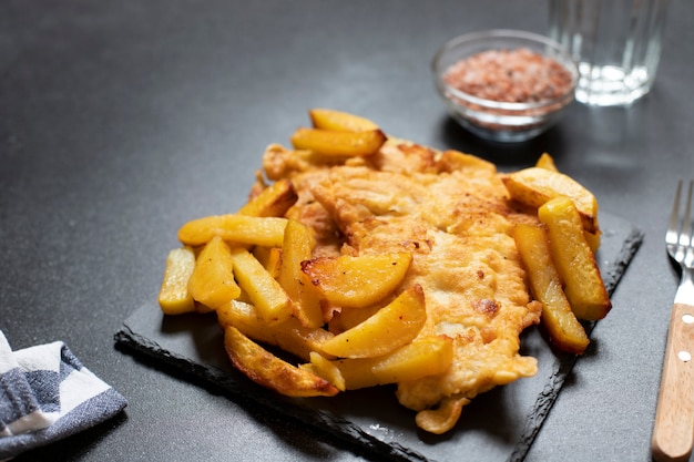 Pescado tradicional británico con patatas fritas. Sobre un plato negro y un fondo negro, servido con sal marina y un vaso de agua. Espacio para texto