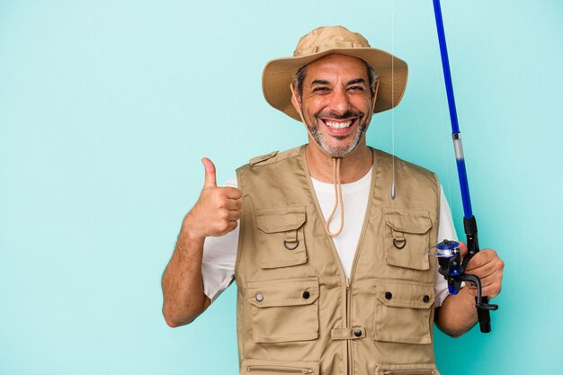 Pescador caucásico de mediana edad sosteniendo la varilla aislado sobre fondo azul sonriendo y levantando el pulgar hacia arriba