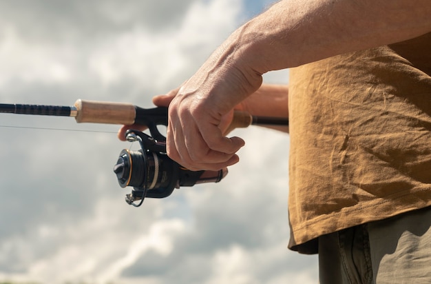 Foto pescador com as mãos segurando a haste do carretel giratório ou isca de colher e pesca de perto. céu de verão com nuvens no fundo.