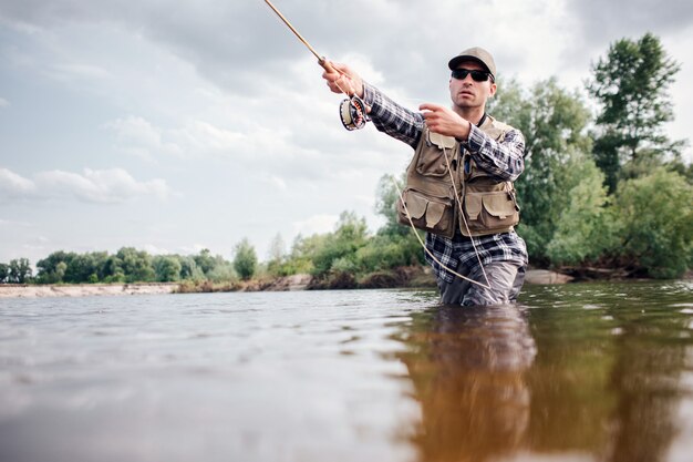 Foto pescador em ação. cara está jogando a colher de cana na água e segurando parte dela na mão. ele olha para a frente. homem usa roupas de proteção especial.