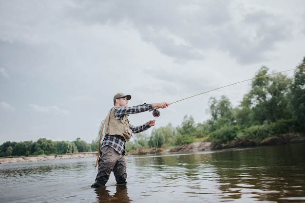 Pescador está parado em águas rasas e pesca. Ele está jogando a colher para a frente. Cara tem haste de mosca nas mãos. Ele está usando isso. Homem usa limícolas, colete e boné para proteger o corpo.