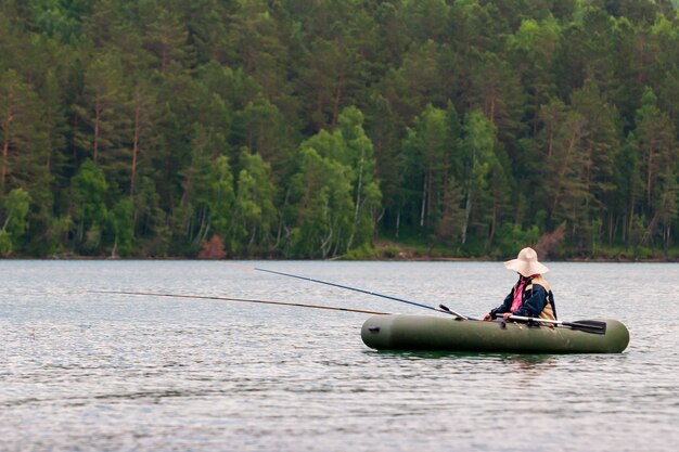 Un pescador está pescando con cañas de pescar desde un bote de goma con remos. El pescador tiene un gran sombrero para el sol. Dos cañas de pescar. Enfoque selectivo. Árboles sobre un fondo borroso.