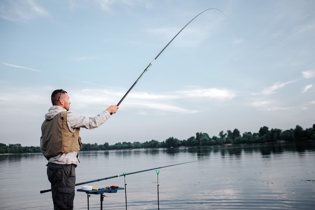 El pescador está de pie cerca del agua y sostiene la caña con la mano derecha. Lo tiene muy alto. El hombre mira hacia adelante. Otra caña de pescado yace en el anzuelo.