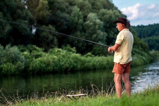 Un pescador con pantalones cortos, un sombrero y una camiseta pesca en la orilla del lago. Pesca, pasatiempos, recreación